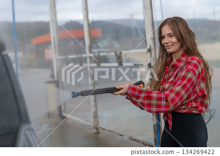 Woman car wash pressure washer uses high stream water to clean black vehicle at self-service station. Woman car wash pressure washer uses high stream water to clean black vehicle at self-service station. 134269422