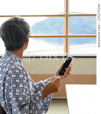 一位身穿浴衣的老人望著窗外的大海，擺弄著他的智慧型手機。 134270164