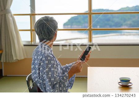 An elderly person in a yukata looks at the view outside the window and their smartphone 134270243