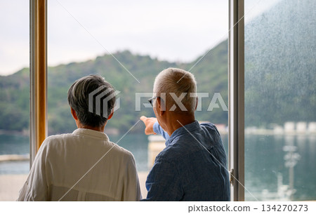 An elderly couple looking out the window at the sea and mountains An elderly couple looking out the window at the sea and mountains 134270273