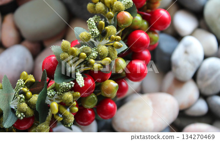 Fragment Of Decorative Chaplet In Branch Form With Colored Berries And Leaves Over Blurred Pebbles Background 134270469
