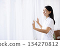 A woman in a white coat smiling and opening the curtains 134270812