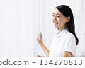 A woman in a white coat smiling and opening the curtains 134270813