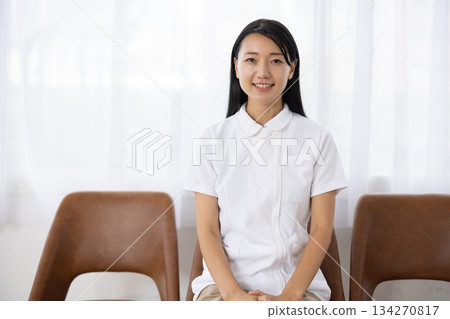 A woman in a white coat sitting in a chair by the window and smiling 134270817