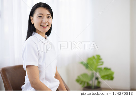 一位身穿白大褂的女士坐在窗邊的椅子上，面帶微笑。 134270818