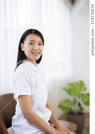 一位身穿白大褂的女士坐在窗邊的椅子上，面帶微笑。 134270819