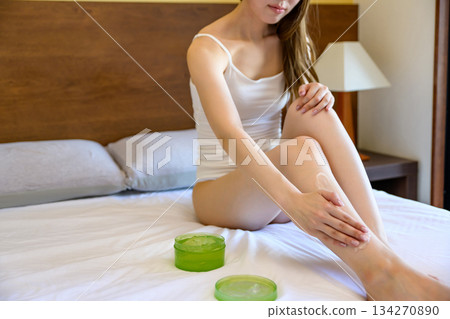 A young, slender woman in underwear applying body gel to her legs in bed. Foot care. Daytime color. 134270890