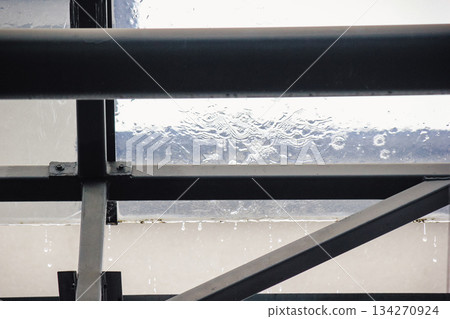Rain falling on a transparent glass roof with water dripping from the dark metal support structure below Rain falling on a transparent glass roof with water dripping from the dark metal support structure below 134270924