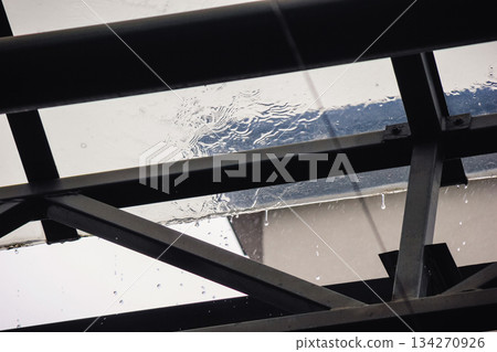 Rain falling on a transparent glass roof with water dripping from the dark metal support structure below Rain falling on a transparent glass roof with water dripping from the dark metal support structure below 134270926