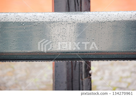 Close up of a smooth, grey metal railing covered in fresh rain drops with a blurred orange background 134270961