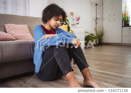 Sad Teenage Girl Sitting on Floor Holding Electronic Cigarette at Home 134271136