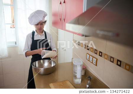 Young Chef In White Hat And Apron Measuring Flour In A Bright Home Kitchen 134271422