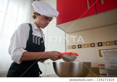 Young Chef In White Hat Sifts Flour Into Mixing Bowl In Modern Kitchen Young Chef In White Hat Sifts Flour Into Mixing Bowl In Modern Kitchen 134271429