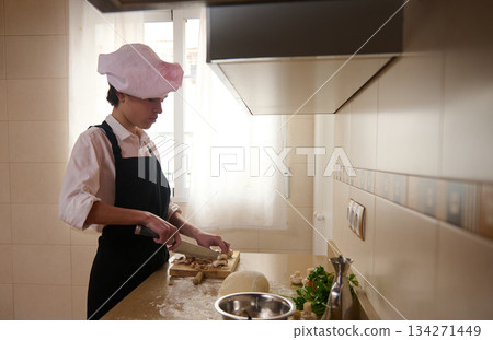 Young Chef In A Pink Toque Chops Mushrooms On A Cutting Board In A Bright Kitchen 134271449