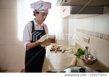 Young Chef In White Shirt And Black Apron Rolling Dough In Bright Kitchen Young Chef In White Shirt And Black Apron Rolling Dough In Bright Kitchen 134271454