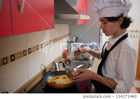 Young Chef In White Uniform And Black Apron Preparing Omelette In Red Modern Kitchen Young Chef In White Uniform And Black Apron Preparing Omelette In Red Modern Kitchen 134271460