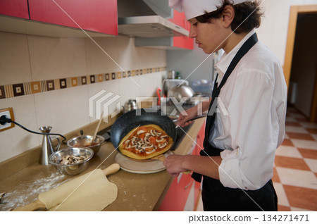 Young Chef In White Shirt And Apron Cooks Pizza In Bright Kitchen Young Chef In White Shirt And Apron Cooks Pizza In Bright Kitchen 134271471