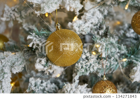 Glittery gold christmas ornament hanging on a flocked christmas tree branch. Holiday decoration for Christmas and New Year celebration. Glittery gold christmas ornament hanging on a flocked christmas tree branch. Holiday decoration for Christmas and New Year celebration. 134271843