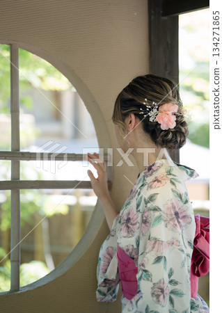A young woman in a yukata robes looks out at the garden from a round window in a teahouse. Kyoto City, Kyoto Prefecture, Japan 134271865