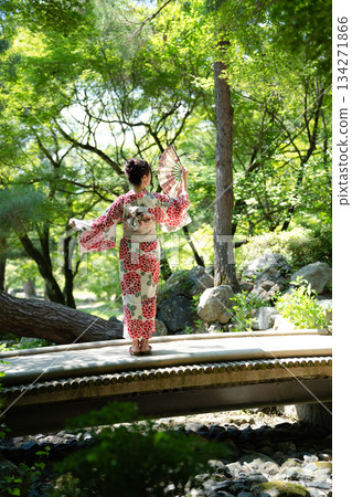 A woman in a yukata holding a folding fan on a bridge in a garden with fresh greenery. Kyoto City, Kyoto Prefecture, Japan 134271866