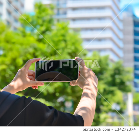 一位女性上半身出現在鏡頭中，她橫握著智慧型手機，在夏日城市拍攝風景影片。 134272374