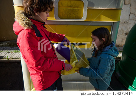 Two Children Recycling Together With Yellow Bag In Front Of Large Recycling Bins 134273414