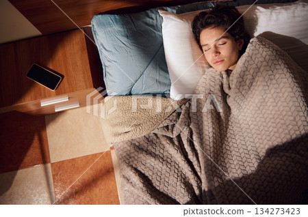 Person Sleeping In Bed With Cozy Blanket, Pillow, And Phone On Nightstand Person Sleeping In Bed With Cozy Blanket, Pillow, And Phone On Nightstand 134273423