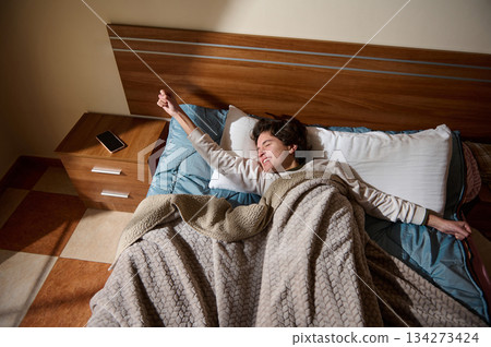 Young Man Laughing And Stretching In Bed With Cozy Blanket In Bedroom Morning 134273424