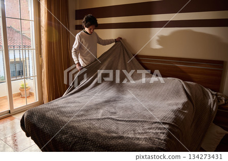 Person Makes Bed In Cozy Bedroom With Blanket, Sunlight, And Striped Wall Decor Person Makes Bed In Cozy Bedroom With Blanket, Sunlight, And Striped Wall Decor 134273431