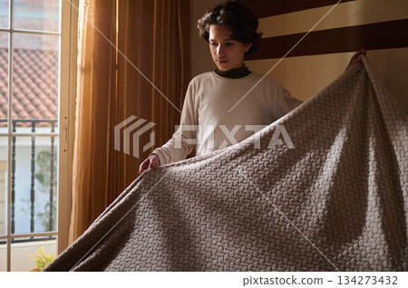 Young Man Unfolds Cozy Blanket In Sunlit Room For Comfort And Warmth Young Man Unfolds Cozy Blanket In Sunlit Room For Comfort And Warmth 134273432