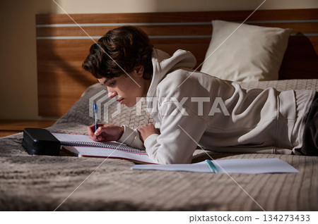 Student Lying on Bed Studying With Notebook and Pen in Cozy Bedroom 134273433