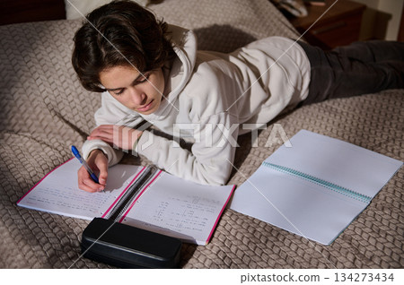 Student Studying at Home Lying on Bed Writing Notes with Notebook and Pencil Case Student Studying at Home Lying on Bed Writing Notes with Notebook and Pencil Case 134273434