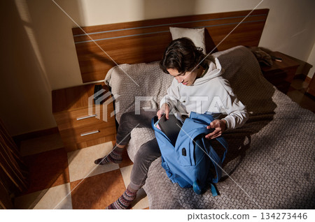 Young Man Organizing Blue Backpack With Notebook on Bed in Cozy Bedroom 134273446
