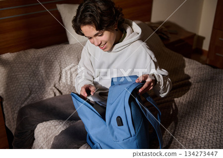 Young Man in White Hoodie Packs Blue Backpack on Bed at Home 134273447