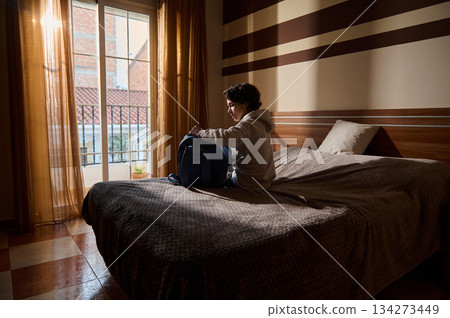 Young Man Packing Backpack on Bed in a Sunlit Hotel Room by a Balcony Window Young Man Packing Backpack on Bed in a Sunlit Hotel Room by a Balcony Window 134273449