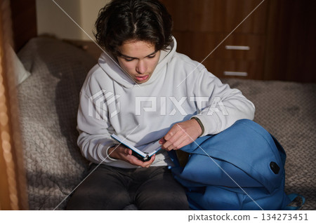 Young Man Reads Passport Handbook at Home With Blue Backpack Nearby Young Man Reads Passport Handbook at Home With Blue Backpack Nearby 134273451