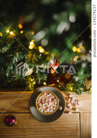 Holiday, Christmas background. A cup with hot chocolate and marshmallows on the table with Christmas tree on background Holiday, Christmas background. A cup with hot chocolate and marshmallows on the table with Christmas tree on background 134274237