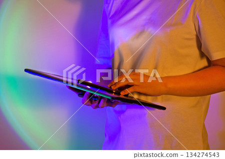 Vibrant work setting and young woman in white shirt with digital tablet in neon bright saturated lights. Power of tech and neon artistry. 134274543