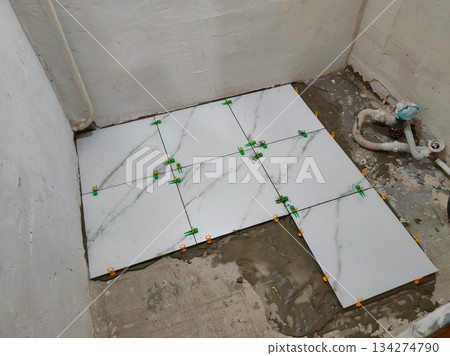 Laying new ceramic floor tiles using a leveling system and spacers on a concrete subfloor. Laying tiles using adhesive during a bathroom renovation, which mirrors the home improvement work. Laying new ceramic floor tiles using a leveling system and spacers on a concrete subfloor. Laying tiles using adhesive during a bathroom renovation, which mirrors the home improvement work. 134274790