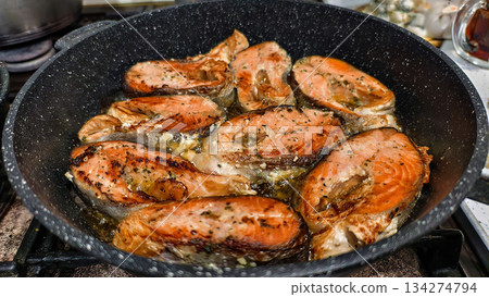 Fried salmon pieces, seasoned with herbs and pepper, sizzle in a frying pan. The golden crust glistens with oil as the pan sits on the stove. A home cook is searing fish fillets for dinner. 134274794