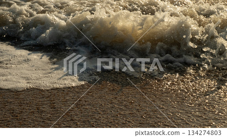 An ocean wave crashes onto wet sand, white sea foam catching golden rays of sunlight and reflecting warm hues. A close-up coastal seascape evokes a sense of calm and natural tranquility. 134274803