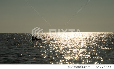 Two people paddleboarding on a calm ocean, silhouetted against the setting sun reflecting brightly and sparkling on the water surface, creating a peaceful and serene scene. 134274833