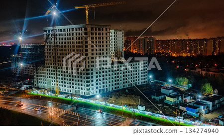 An urban construction site. Several tower cranes illuminate the unfinished concrete structures, while traffic moves along the wet road below. A new building is under construction. 134274940