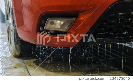 A wet car bumper with dripping water, shiny orange paint, and headlights. A car wash with pressure water creates dynamic splashes and soap streaks. A close-up of the car's bumper at the car wash. A wet car bumper with dripping water, shiny orange paint, and headlights. A car wash with pressure water creates dynamic splashes and soap streaks. A close-up of the car's bumper at the car wash. 134275040
