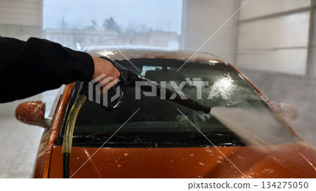 A man washes a car windshield in a covered garage with high-pressure water and foam. His hand holds the sprayer as he washes the car, creating steam and droplets on the glass. A man washes a car windshield in a covered garage with high-pressure water and foam. His hand holds the sprayer as he washes the car, creating steam and droplets on the glass. 134275050