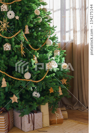 Festive Christmas Tree with natural wooden ornaments and garland with wrapped holiday gifts boxes Festive Christmas Tree with natural wooden ornaments and garland with wrapped holiday gifts boxes 134275144