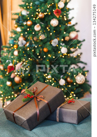 Two Christmas gift boxes decorated with ribbon and red holly berry in front of glowing festive tree 134275149
