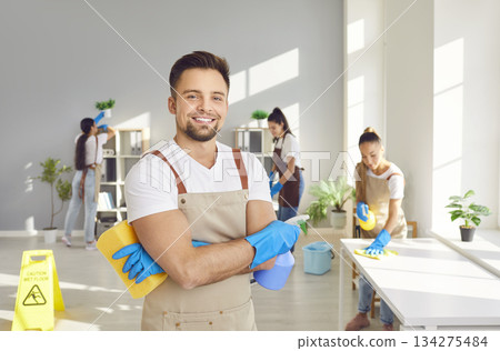 Happy male young janitor in uniform looking at camera holding household cleaning supplies Happy male young janitor in uniform looking at camera holding household cleaning supplies 134275484