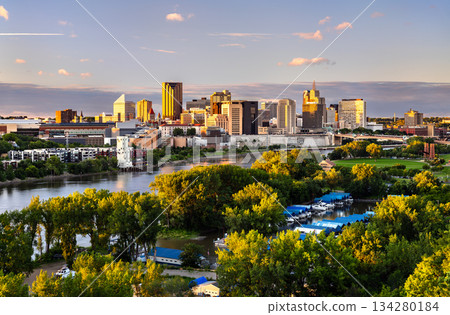 Downtown Saint Paul skyline rises above the Mississippi River in Minnesota, USA. Scenic view features urban architecture and green trees under a sunset sky 134280184