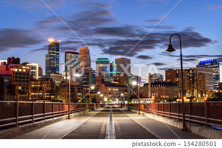 Night view from Stone Arch Bridge features Minneapolis skyline and Mississippi River. Illuminated walking path leads to downtown skyscrapers under blue sky Night view from Stone Arch Bridge features Minneapolis skyline and Mississippi River. Illuminated walking path leads to downtown skyscrapers under blue sky 134280501
